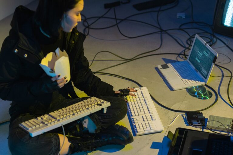 Top 10 Cybersecurity Threats for IT Teams in 2026 A young woman in a dark room surrounded by computers and cables, eating and typing on keyboards.