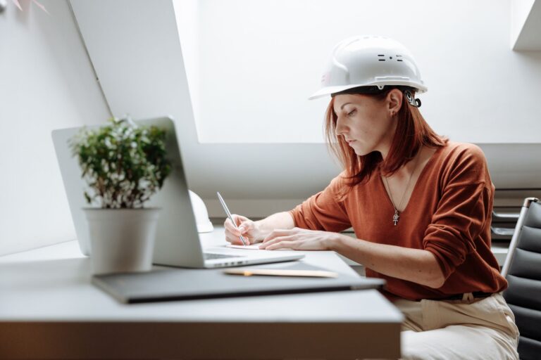 AWS Solutions Architect Associate: Complete Study Guide A focused female architect in a hard hat writing notes while working on a laptop indoors.
